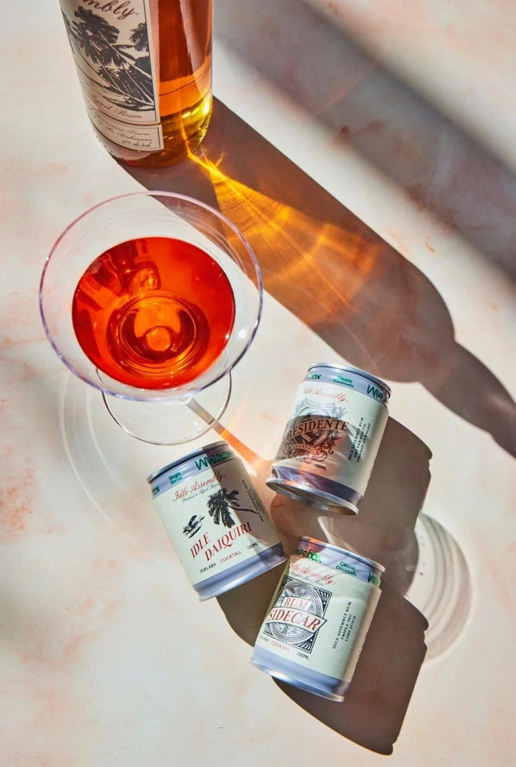 Idle Assembly cocktail cans and a bottle beside a cocktail glass in bright sunlight